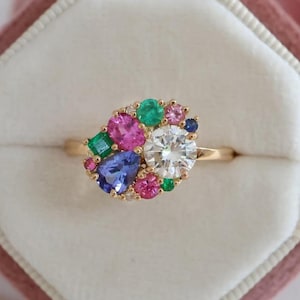 May include: A gold ring featuring a cluster of colorful gemstones. The ring has a large, round, clear center stone surrounded by pink, green, blue, and other colored gems. The ring is set in a gold band and is displayed in a pink velvet ring box.