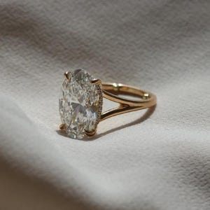 May include: A gold ring with a large, oval-cut diamond as the centerpiece. The diamond is clear and sparkling, set in a gold band with a split shank design. The ring is displayed on a white fabric surface.