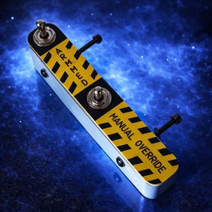 Puede incluir: Un dispositivo metálico rectangular con un diseño de rayas amarillas y negras. El texto "ARMED" y "MANUAL OVERRIDE" están impresos en el dispositivo. Tiene dos interruptores de palanca y dos tornillos negros. El fondo es una galaxia azul.