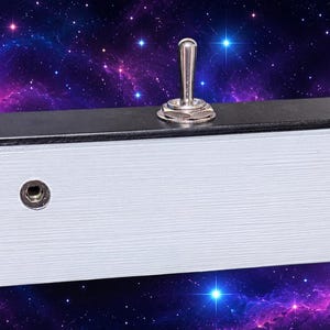 Könnte beinhalten: Ein weißes rechteckiges Gerät mit zwei silbernen Kippschaltern und zwei kreisförmigen Buchsen. Die Oberseite ist schwarz. Das Gerät befindet sich vor einem Hintergrund aus einer lila und blauen Galaxie.