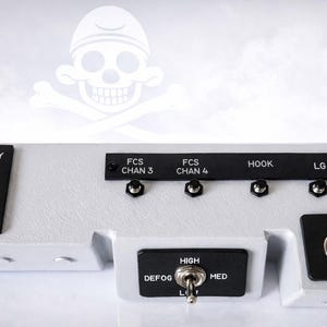 Pode incluir: Um painel de controle cinza com etiquetas e interruptores pretos. O texto inclui "CANOPY", "OPEN", "HOLD", "CLOSE", "FCS CHAN 3", "FCS CHAN 4", "HOOK", "LG", "DEFOG", "HIGH", "MED", "ON" e "FCALIT". Um gráfico de caveira e ossos cruzados está no fundo.