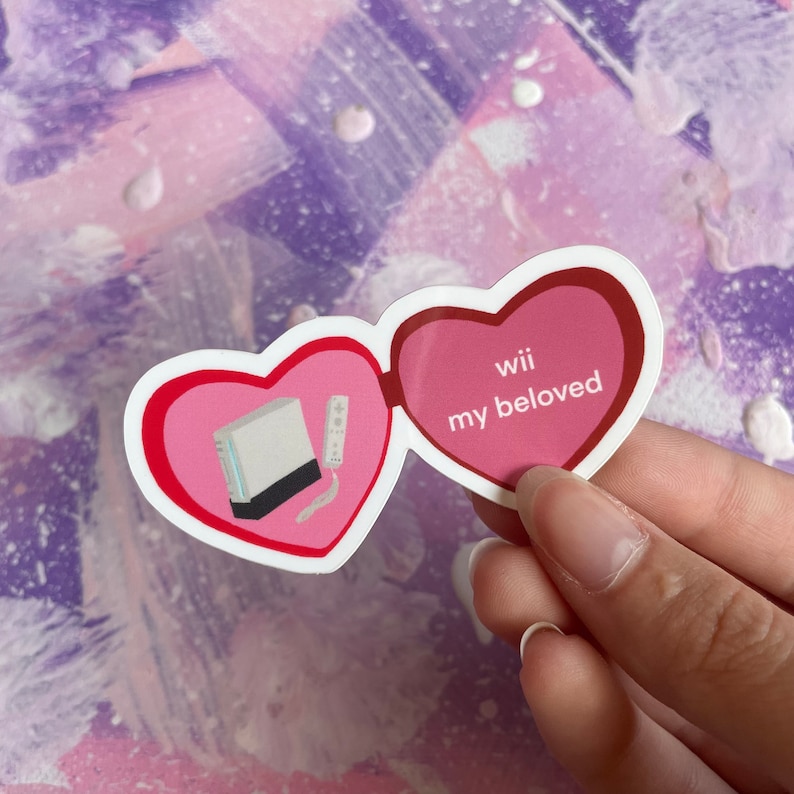 May include: A pink and red heart-shaped sticker with a white Nintendo Wii console and a remote inside one heart and the text "Wii my beloved" inside the other heart.