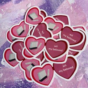 May include: A stack of pink heart-shaped stickers with a white Nintendo Wii console and remote printed on them. The stickers have the text "Wii my beloved" printed on them.