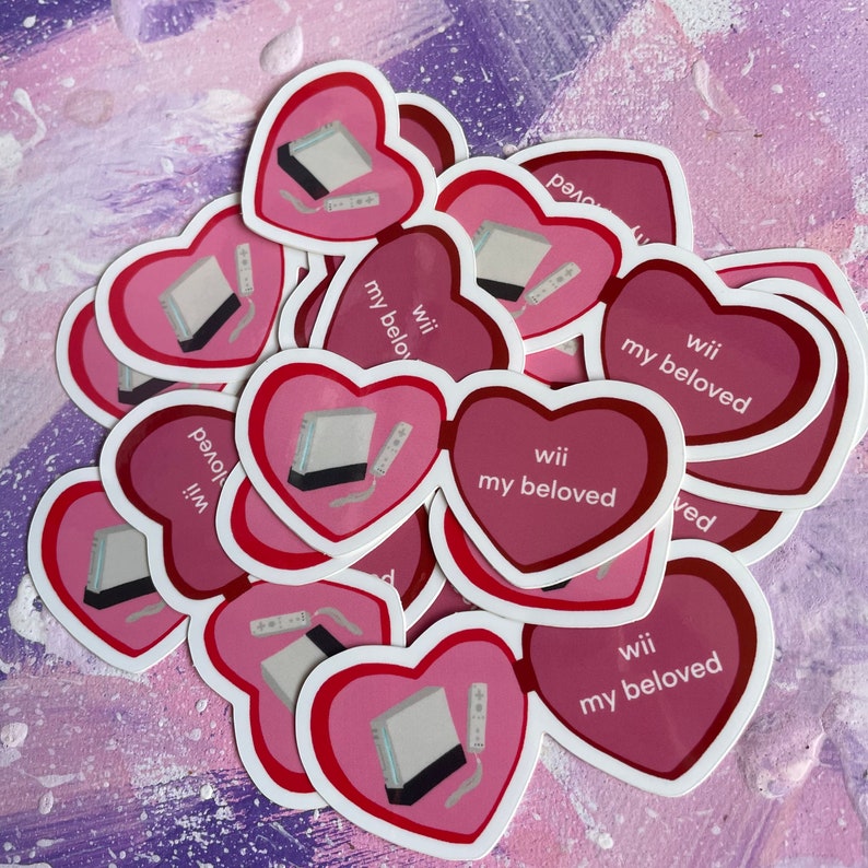 May include: A stack of pink heart-shaped stickers with a white Nintendo Wii console and remote printed on them. The stickers have the text "Wii my beloved" printed on them.