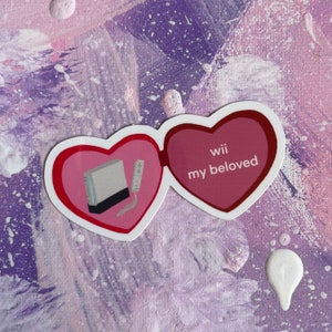 May include: A pink heart-shaped sticker with a white Nintendo Wii console and a white remote inside. The other heart says "Wii my beloved".