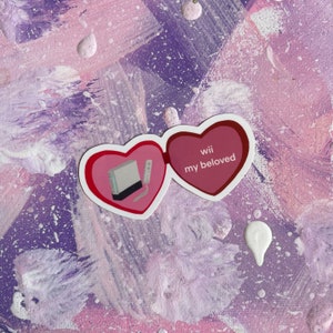 May include: A pink and red heart-shaped sticker with a white Nintendo Wii console and a remote control on the left side and the text "Wii my beloved" on the right side.