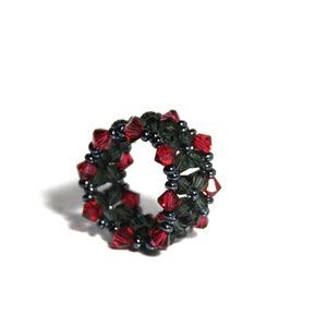 Könnte beinhalten: Ein Perlenring mit einem sich wiederholenden Muster aus dunkelgrünen und roten Kristallen. Der Ring besteht aus kleinen, dunklen Perlen und hat ein kreisförmiges Design. Die Kristalle sind facettiert und reflektieren das Licht. Der Ring befindet sich auf weißem Hintergrund.
