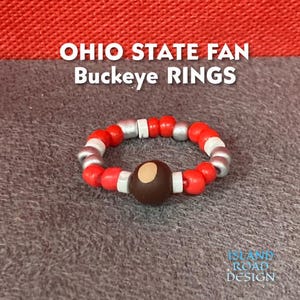 May include: A red, white, and silver beaded ring with a brown buckeye nut charm. The ring is on a gray background. OHIO STATE FAN Buckeye RINGS is written in white text on a red background.