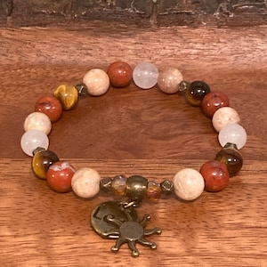 May include: A brown, white, and red beaded bracelet with a gold charm in the shape of a sun. The bracelet is on a wooden surface.