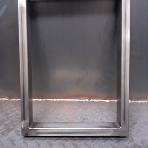 May include: A silver metal frame with a rectangular shape. The frame is made of thick metal and has a welded construction. The frame is sitting on a gray textured surface.