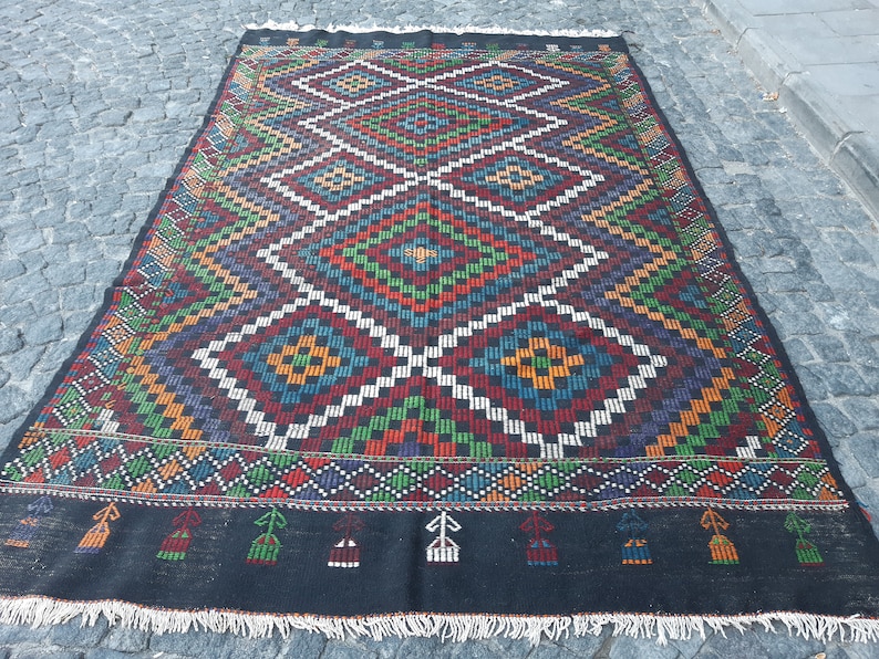 May include: A colourful, hand-woven rug with a geometric pattern in shades of red, blue, green, yellow, and white. The rug has a black background and a fringe border.