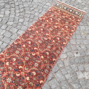 May include: A red, blue, and cream patterned runner rug with a floral design. The rug is laid out on a gray cobblestone surface.