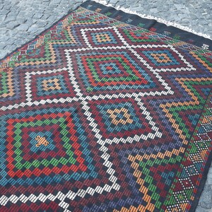 May include: A colourful rug with a geometric pattern in shades of red, green, blue, orange, and white. The rug has a black background and a fringe border.