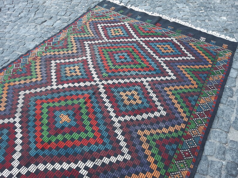 May include: A colourful rug with a geometric pattern in shades of red, green, blue, orange, and white. The rug has a black background and a fringe border.
