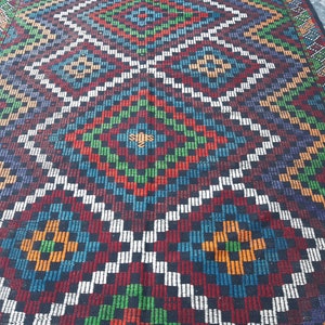 May include: A colourful woven rug with a geometric pattern in shades of red, green, blue, white, and black. The rug has a diamond-shaped design with a central motif.