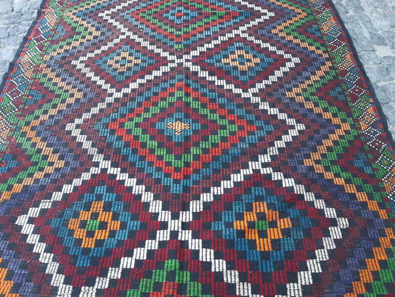 May include: A colourful woven rug with a geometric pattern in shades of red, green, blue, white, and black. The rug has a diamond-shaped design with a central motif.