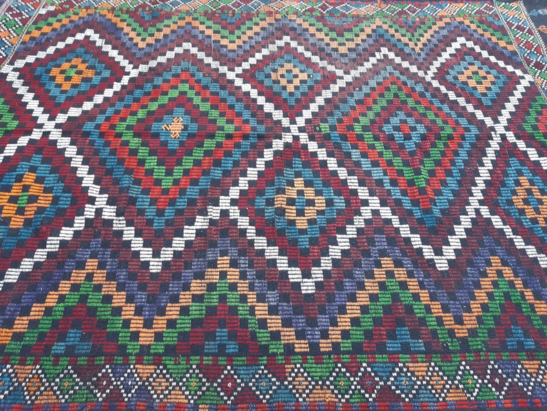 May include: A colourful rug with a geometric pattern in shades of red, green, blue, white, and black. The rug has a diamond pattern with smaller squares within the diamonds.