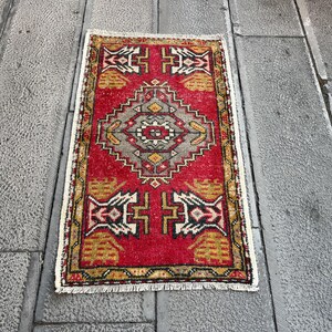 May include: A red, gold and white patterned rug with a fringed edge. The rug has a traditional design with geometric shapes and floral motifs.