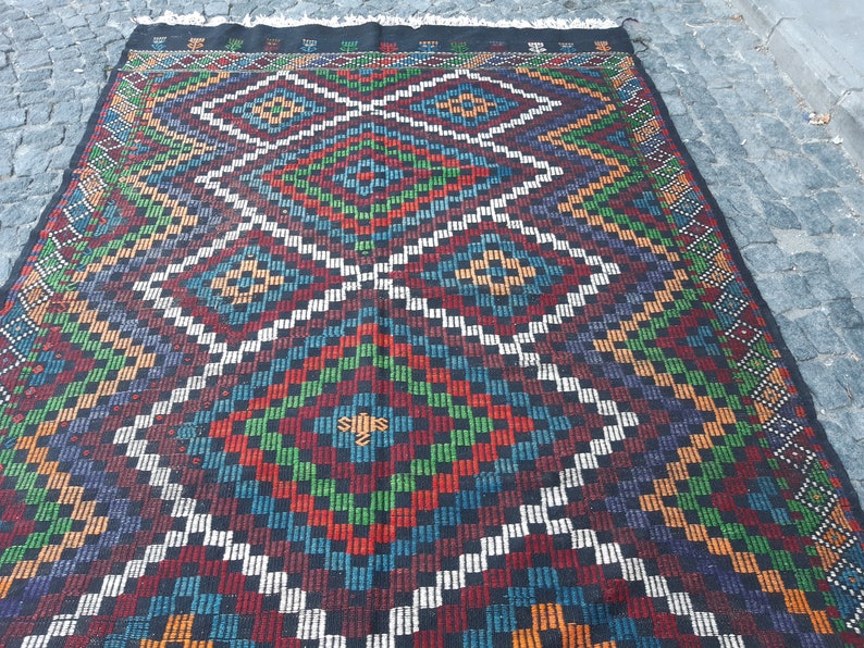 May include: A colourful rug with a geometric pattern in shades of red, green, blue, white, and black. The rug has a diamond-shaped design with intricate details.