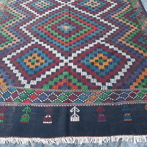 May include: A colourful handwoven rug with a geometric pattern in shades of red, green, blue, white, and black. The rug has a fringe border.