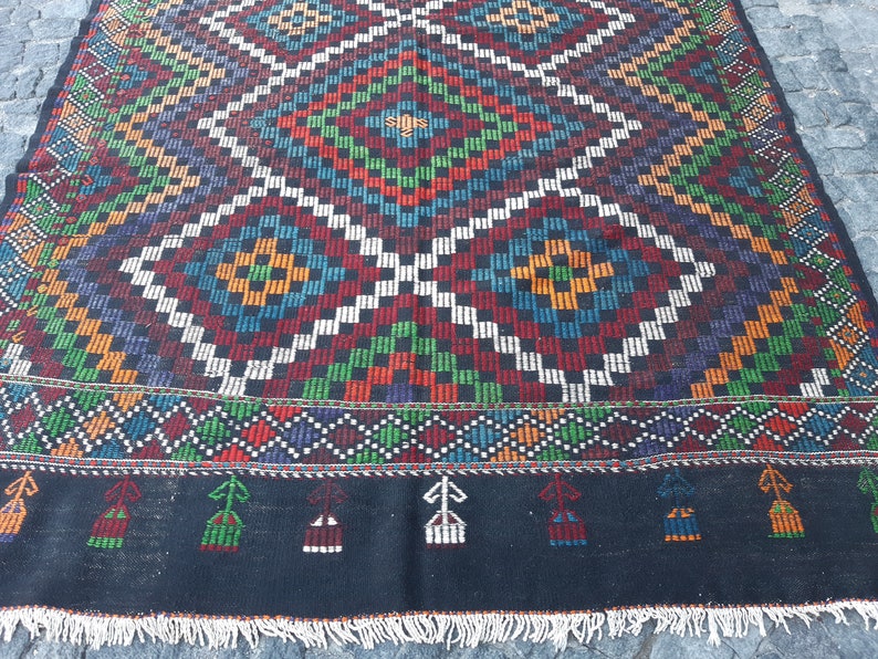 May include: A colourful handwoven rug with a geometric pattern in shades of red, green, blue, white, and black. The rug has a fringe border.