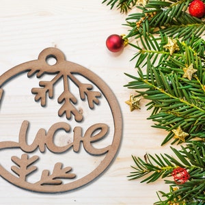 Peut inclure: Une décoration de Noël en bois en forme de cercle avec le nom "Lucie" découpé au centre. La décoration est ornée d'un flocon de neige et de petites branches de pin.
