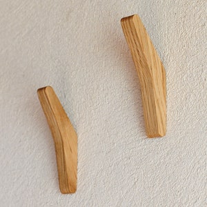 May include: Two wooden wall hooks in a light brown color, mounted on a white wall. The hooks are curved and have a simple, modern design.