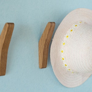 May include: Two wooden wall hooks with a curved design, mounted on a light blue wall. A white straw hat with a yellow daisy trim is hanging on one of the hooks.