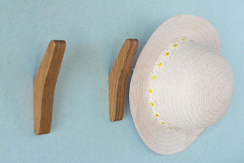May include: Two wooden wall hooks with a curved design, mounted on a light blue wall. A white straw hat with a yellow daisy trim is hanging on one of the hooks.
