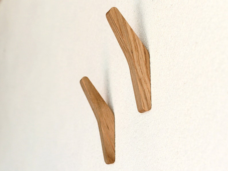 May include: Two wooden wall hooks in a light brown color, mounted on a white wall. The hooks are shaped like a curved, angled line.