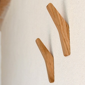 May include: Two wooden wall hooks in a light brown color, mounted on a white wall. The hooks are angled and have a curved shape.