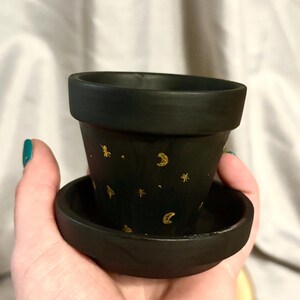 May include: A small, black ceramic plant pot with a matching saucer. The pot is adorned with gold-colored stars and crescent moons. The pot is held in a hand, and the background is a soft, neutral fabric.
