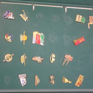 Peut inclure: Une collection d'épingles émaillées colorées exposées sur un fond vert. Les épingles présentent divers motifs, dont un palmier, un avion et le texte "90's". Les épingles sont fixées à un classeur avec le texte "LES PIN'S".