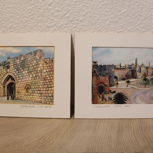 Può includere: Due stampe ad acquerello incorniciate raffiguranti le porte della città di Gerusalemme. La stampa a sinistra mostra la Porta di Sion con mura in pietra e un ingresso ad arco. Quella a destra illustra la Porta di Giaffa con mura, palme e una strada. Entrambe presentano scritte a mano.