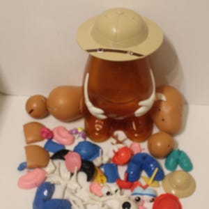 Puede incluir: Una patata de juguete de plástico marrón con varias piezas desmontables, incluyendo brazos, piernas, sombreros y rasgos faciales. El juguete está parcialmente montado, con un sombrero de safari beige en la parte superior. Una colección de accesorios coloridos está dispersa.