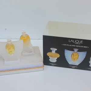 Peut inclure: Un ensemble de flacons de parfum miniatures dans une boîte blanche avec une bordure dorée et une boîte noire avec le texte "LALIQUE PARIS". Les flacons sont transparents avec du liquide jaune à l'intérieur. L'ensemble comprend trois flacons sur une base blanche.