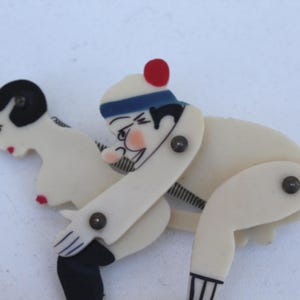 Può includere: Una spilla vintage articolata raffigurante due figure in una posa giocosa. Le figure sono principalmente bianco sporco con accenti neri e rossi. La spilla ha uno stile stravagante e fumettistico, con rivetti visibili sulle articolazioni.
