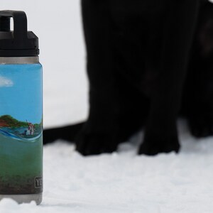 May include: A stainless steel water bottle with a black handle and lid. The bottle features a painted landscape scene with a blue sky, water, and a small boat. The bottle is set against a snowy background, with a black dog in the background.