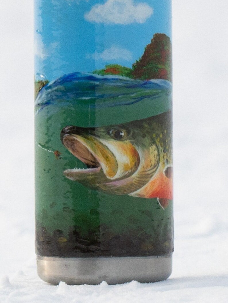 May include: A stainless steel tumbler with a detailed illustration of a fish underwater. The design includes a blue sky, green water, and a fish with orange and brown accents. The tumbler has a silver base.