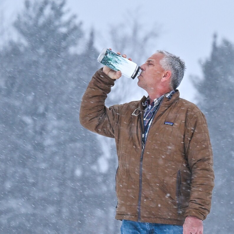 May include: A person drinking from a white and silver water bottle with a forest design. The person is wearing a brown jacket in a snowy environment. The water bottle is cylindrical.
