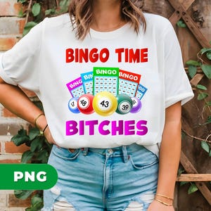 Puede incluir: Camiseta blanca con las palabras "BINGO TIME BITCHES" en rojo y rosa. La camiseta presenta bolas y cartones de bingo en varios colores. La camiseta se combina con pantalones cortos de mezclilla.