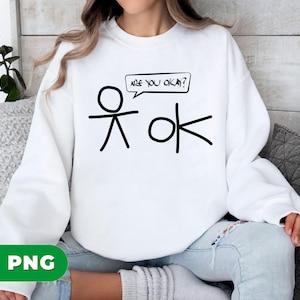 Pode incluir: Camisola branca com um desenho de boneco de palito preto. O desenho inclui um boneco de palito e a palavra "ok" com o texto "ARE YOU OKAY?" numa bolha de fala. A camisola é de estilo casual.