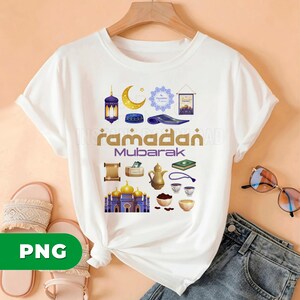 May include: White t-shirt featuring a colorful graphic design with the words "Ramadan Mubarak" and various Islamic symbols. The design includes a crescent moon, mosque, lantern, and prayer rug. The shirt is displayed on a hanger.