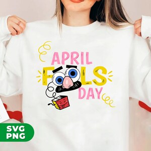 以下が含まれることがあります： カラフルな「エイプリルフールズデー」のグラフィックがプリントされた白いスウェットシャツ。大きな青い目、口ひげ、舌が出ている赤い箱が特徴の漫画の顔のデザイン。テキストはピンクと黄色です。