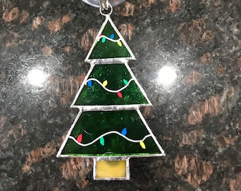 May include: A green stained glass Christmas tree ornament with white lights and colorful bulbs. The ornament has a suction cup on the back for hanging.