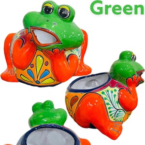 May include: A green frog planter with orange, yellow, blue, and white painted details. The frog is sitting on its hind legs with its front legs crossed. The planter has a wide opening for planting.
