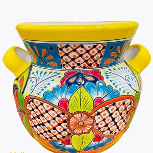 May include: A yellow ceramic planter with a colorful floral design. The planter has two handles on the sides and a wide opening at the top.