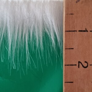 May include: White faux fur trim measuring approximately 7.6 cm in length. The fur is soft and fluffy, and it is shown against a green background.