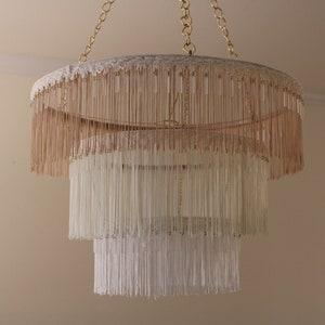 May include: A three-tiered chandelier with a white and beige fringe. The chandelier is made of a woven material and has a circular shape.