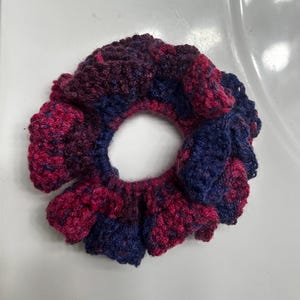 May include: A handmade scrunchie in shades of deep red, purple, and navy blue. The scrunchie is crocheted with a textured stitch, creating a ruffled effect around the elastic band. This hair accessory is perfect for adding a pop of colour to any hairstyle.